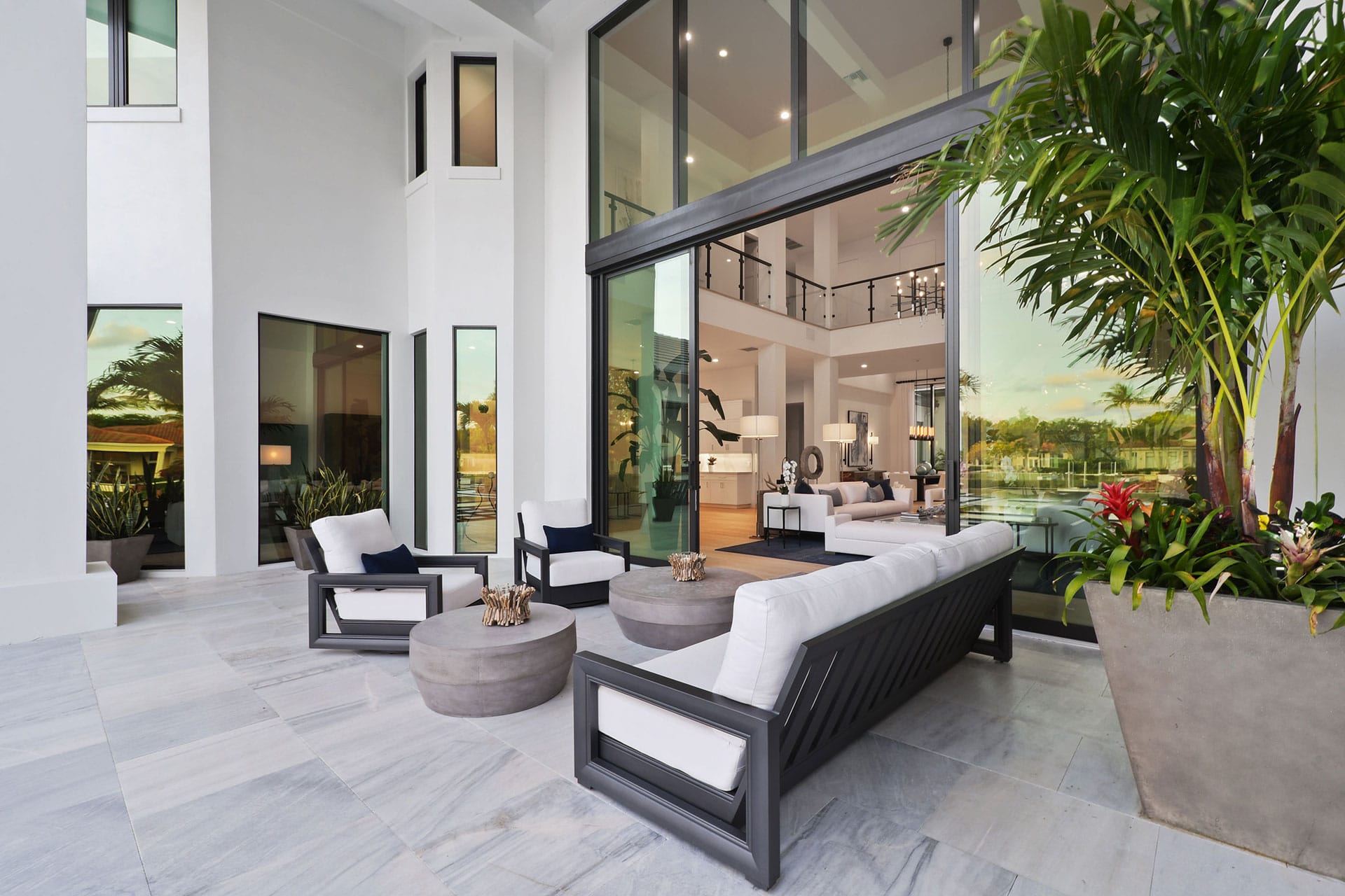 Modern outdoor patio with white cushioned seating, round tables, and large potted plants on elegant stone flooring, opening to a spacious, luxurious interior with high ceilings and glass doors, overlooking water and greenery.