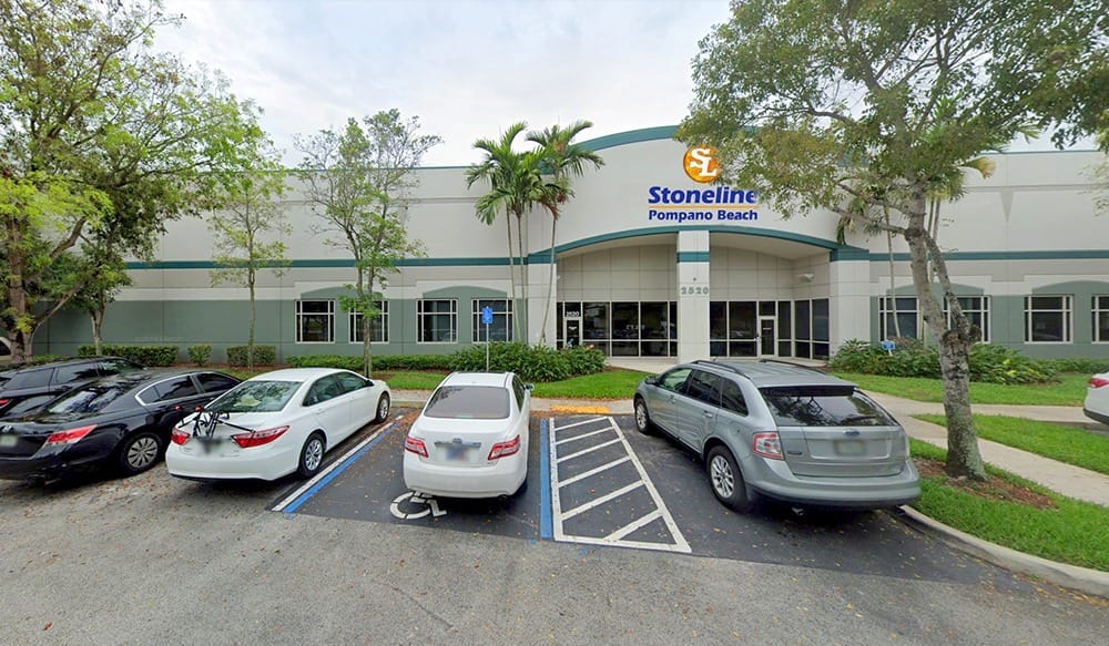 Front view of a commercial building with a sign that reads Stoneline Pompano Beach, several parked cars—including one in a handicapped space—and stone landscaping features with trees and grass surrounding the parking lot.