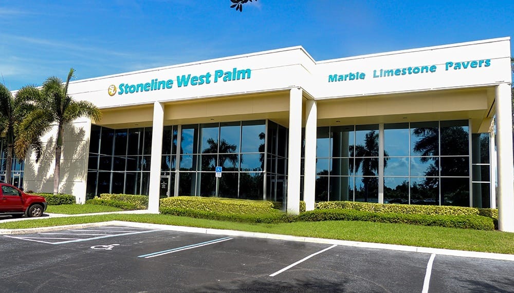 A modern commercial building with large windows, labeled Stoneline West Palm and Marble Limestone Pavers, features elegant stone accents, surrounded by palm trees and a parking lot under a blue sky.